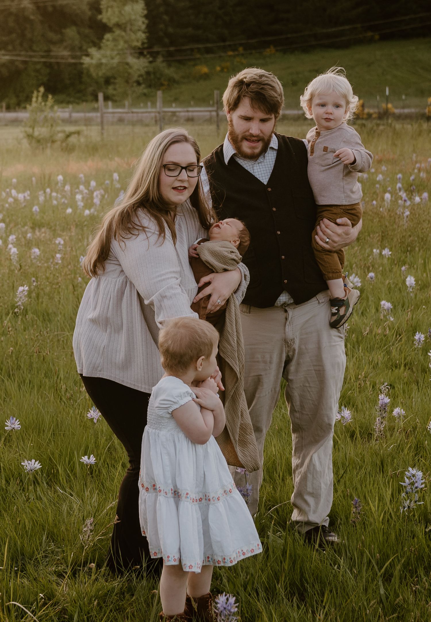 Family of five posing in a wildflower meadow at golden hour, parents holding children, wearing casual elegant attire.
