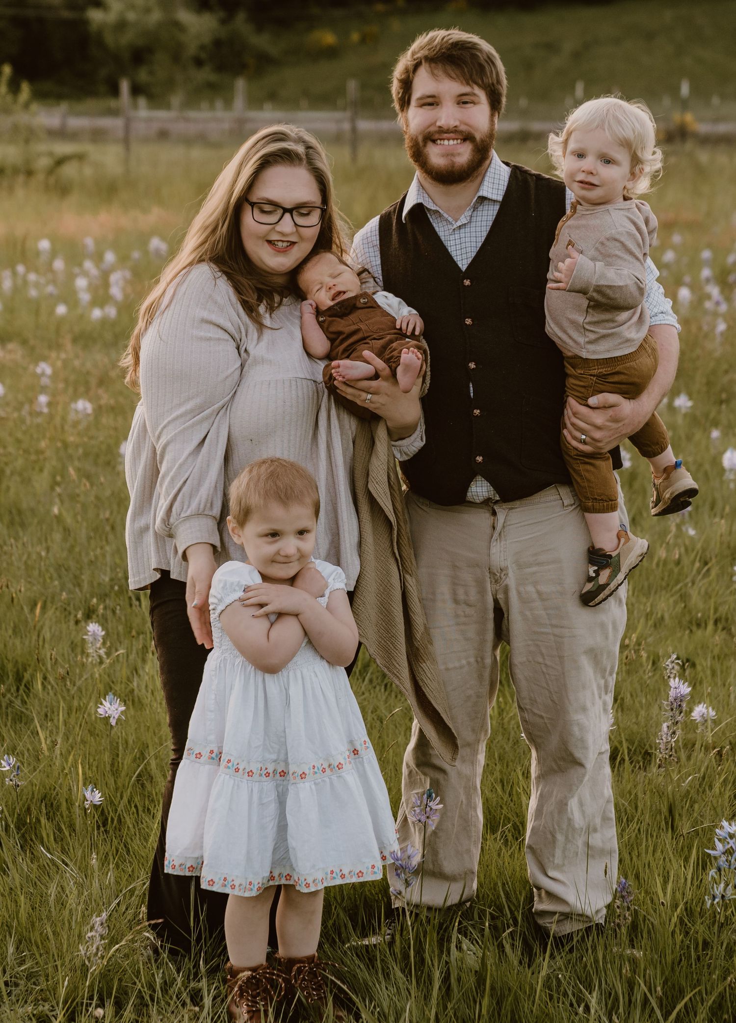 Happy family of five posing in a wildflower meadow at golden hour, parents holding young children outdoors.