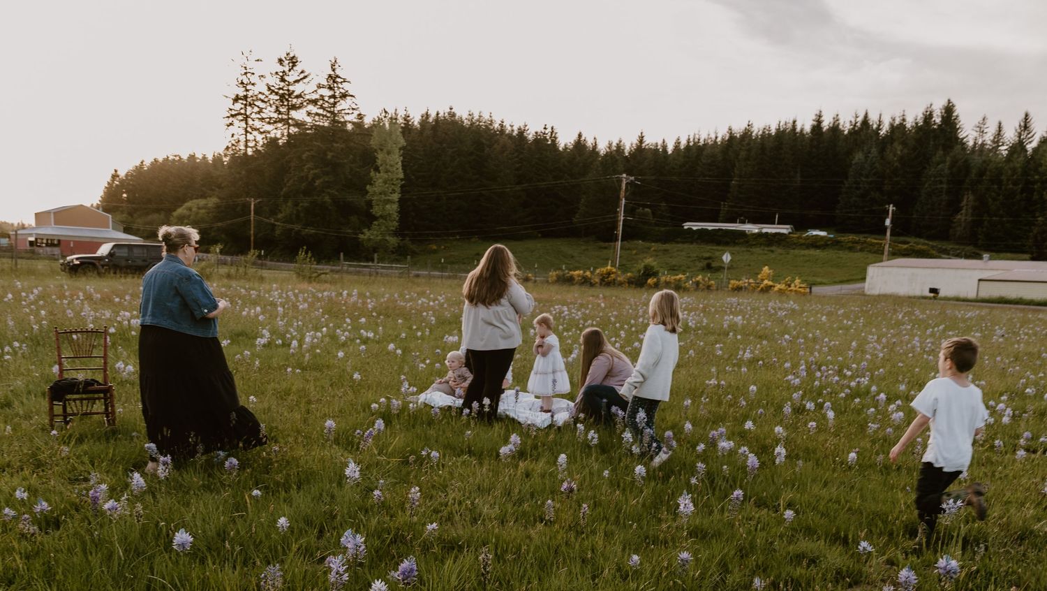 Family gathering in a wildflower meadow at golden hour with children playing among white flowers and tall evergreen trees.