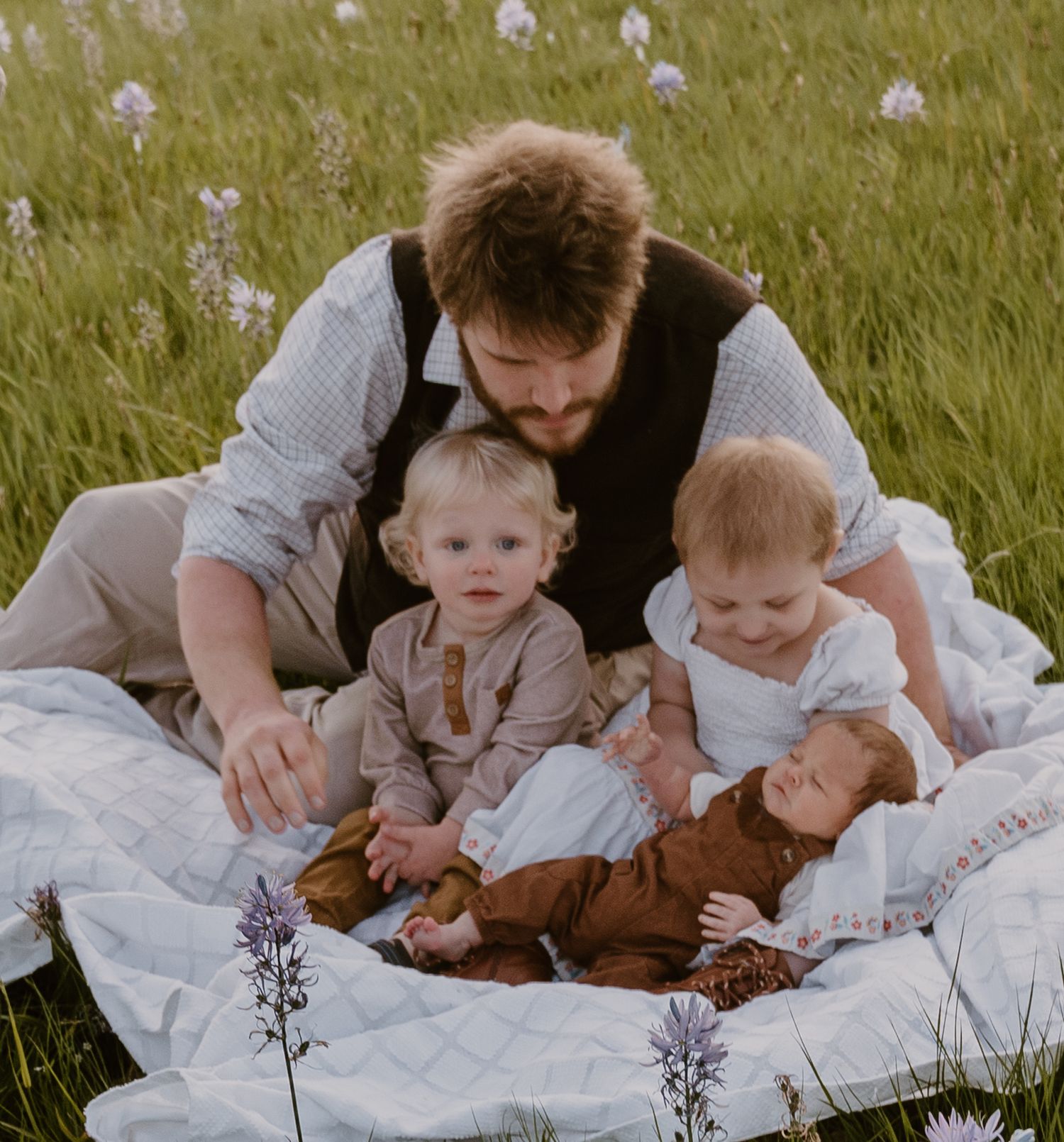 A bearded father sits in a green meadow with three young children, including a newborn, on a white quilt.