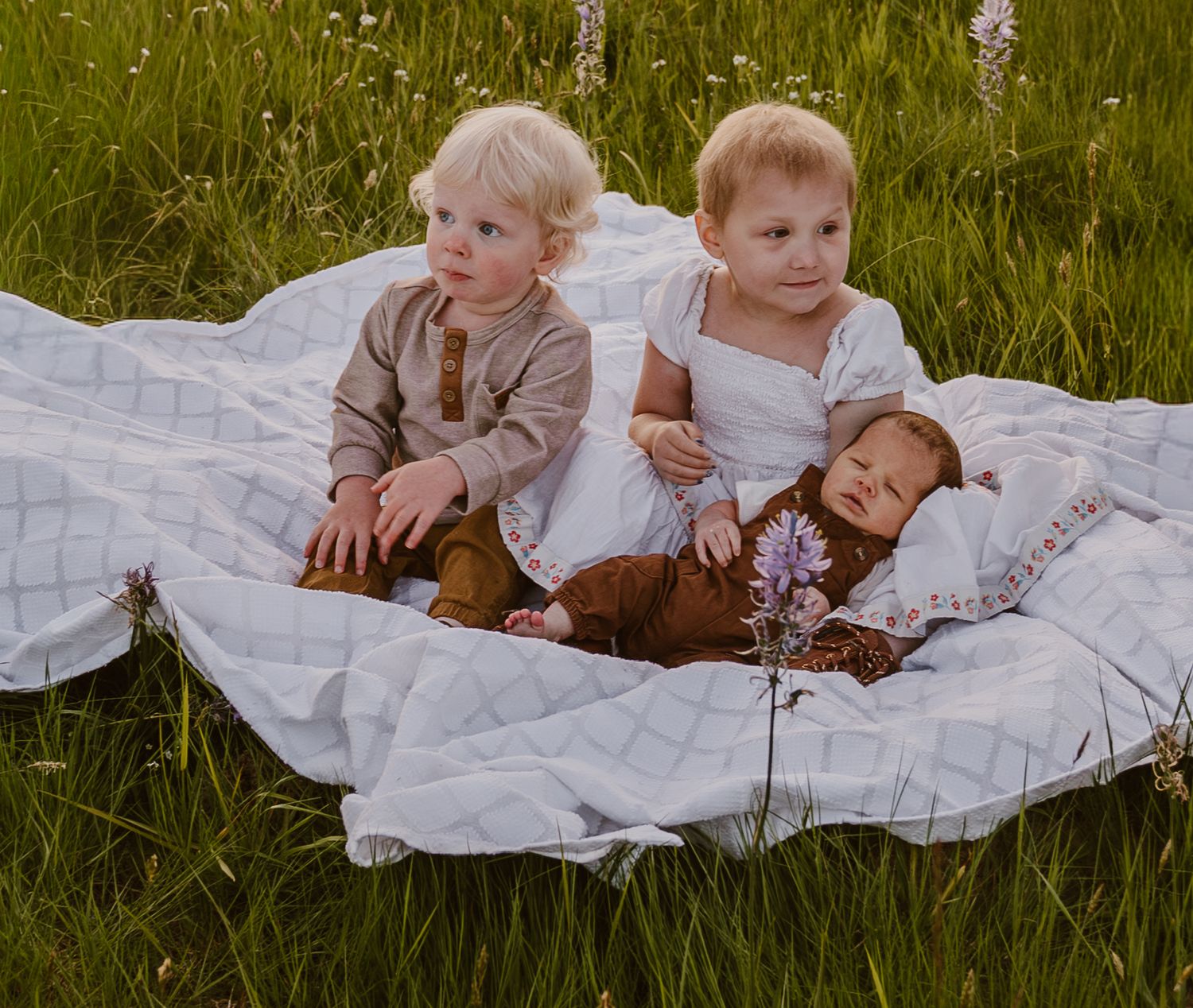 Two toddlers and a newborn baby rest on a white quilt blanket in a lush green grass field.