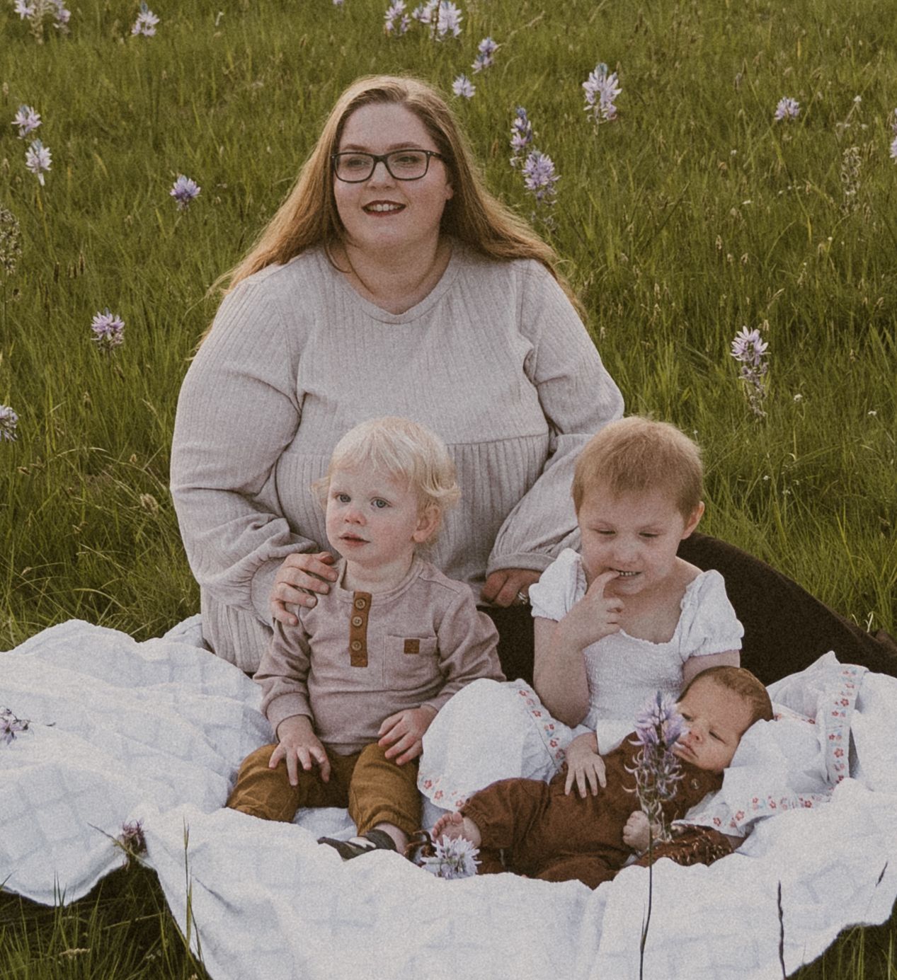 A woman with glasses sits on a blanket in a flower field with two toddlers and a newborn baby.