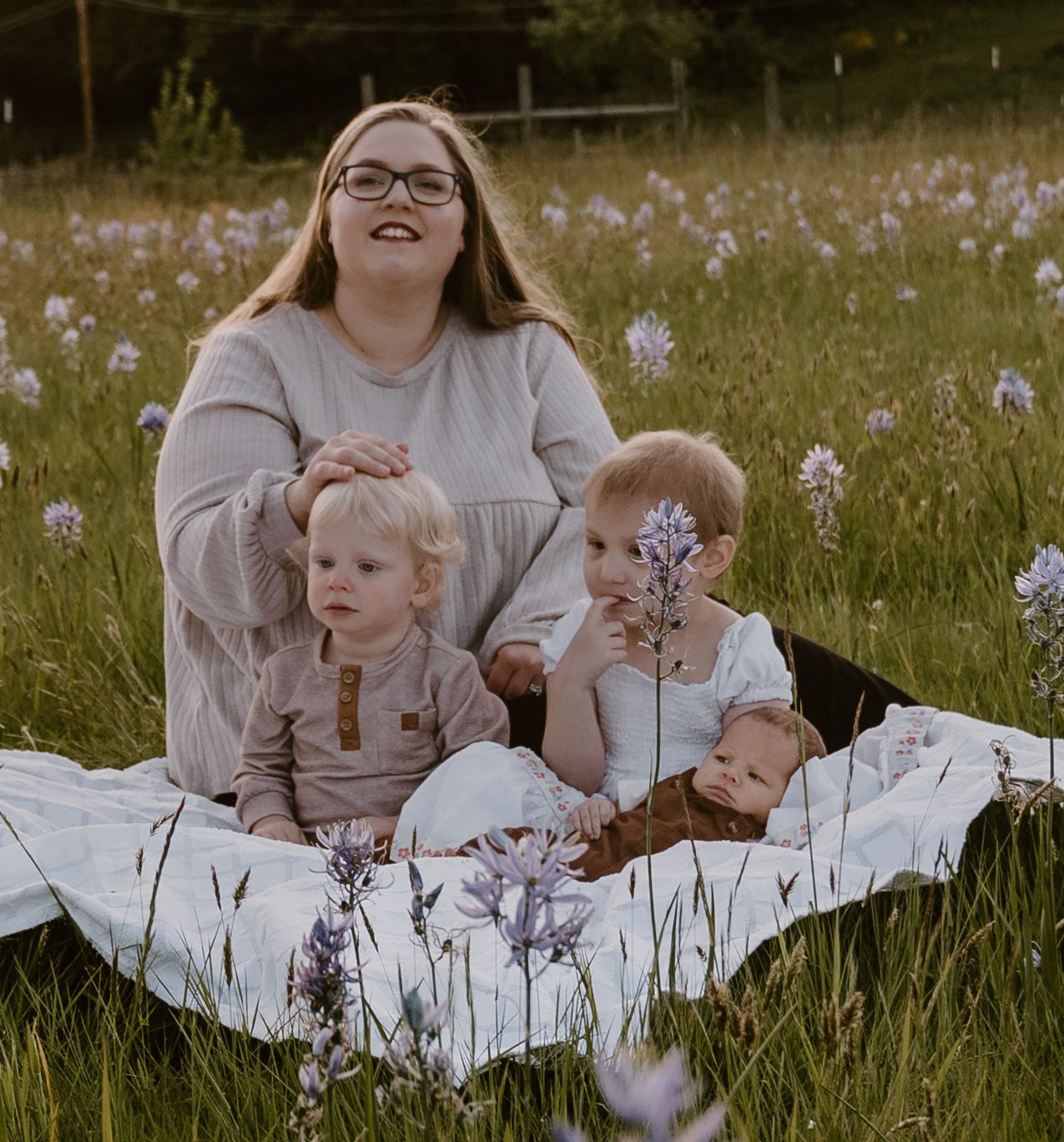 A woman with glasses sits on a white blanket in a wildflower meadow with three young children on a sunny day.
