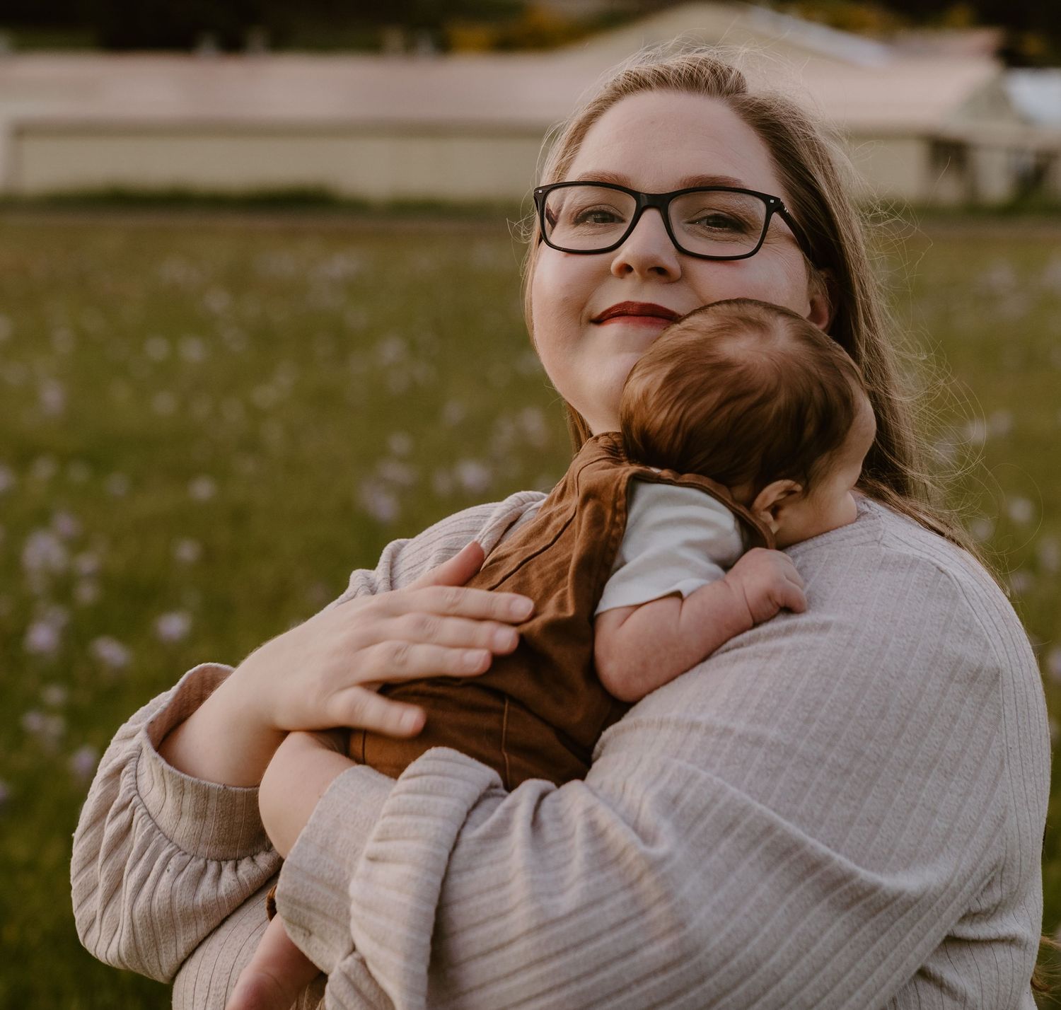 A young woman wearing glasses tenderly holds a sleeping newborn baby outdoors in a green field.