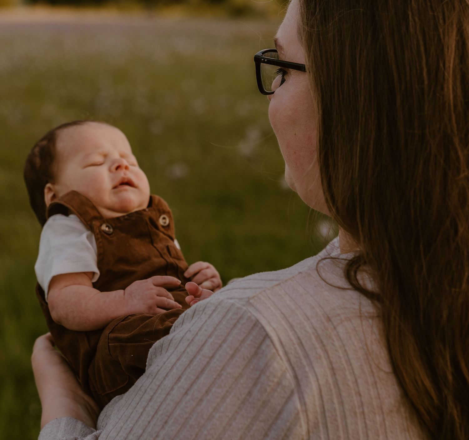 A mother holding her sleeping newborn baby dressed in a brown outfit in a sunlit field.