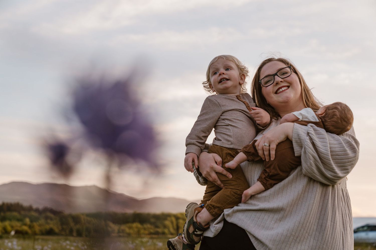 A smiling mother holding a toddler and baby outdoors with purple smoke and mountains in the background.