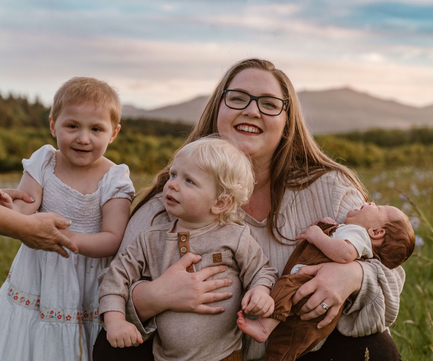 A smiling mother holds three young children in a scenic mountain meadow at sunset.