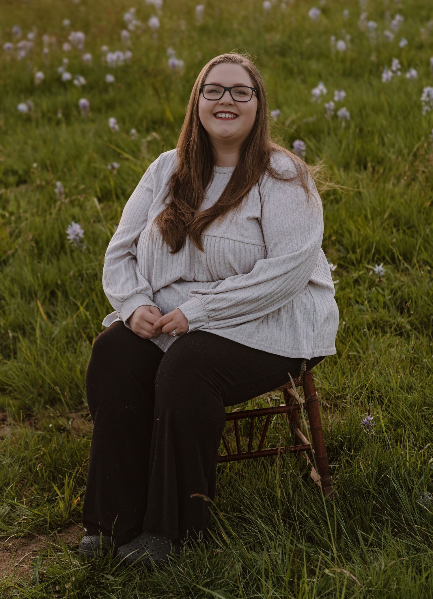 Young woman with glasses sitting on a wooden chair in a green meadow with wildflowers, wearing a white blouse.