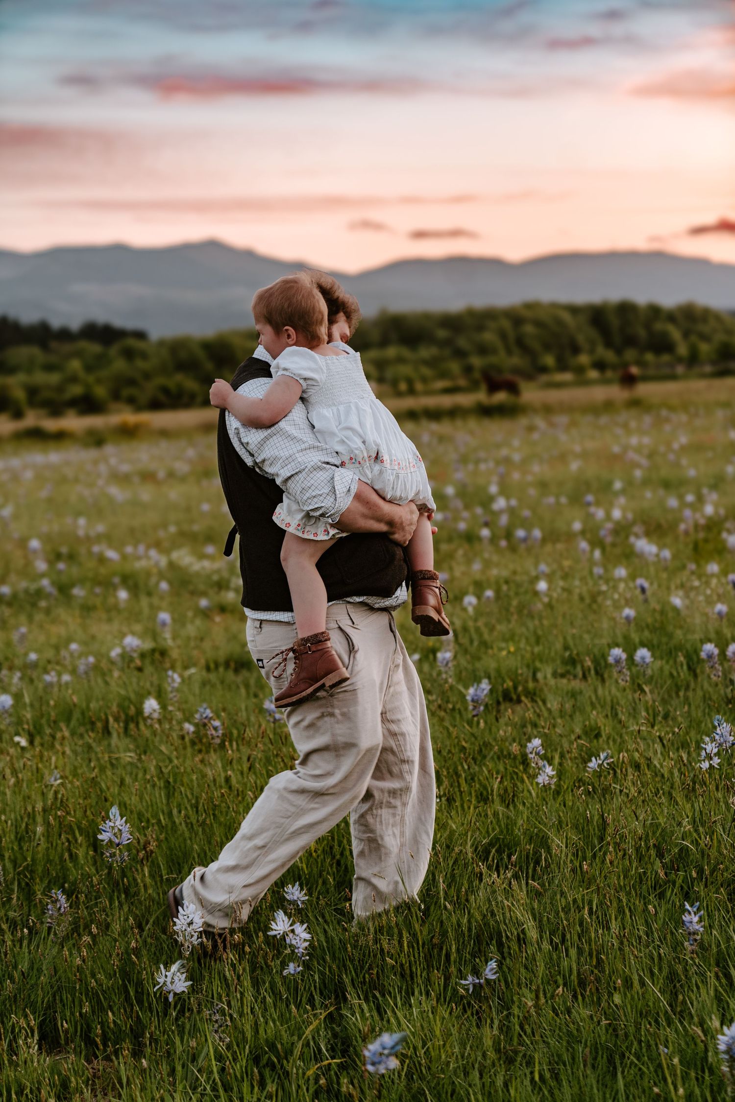 A father carries his toddler through a wildflower meadow at sunset with mountains in the background.