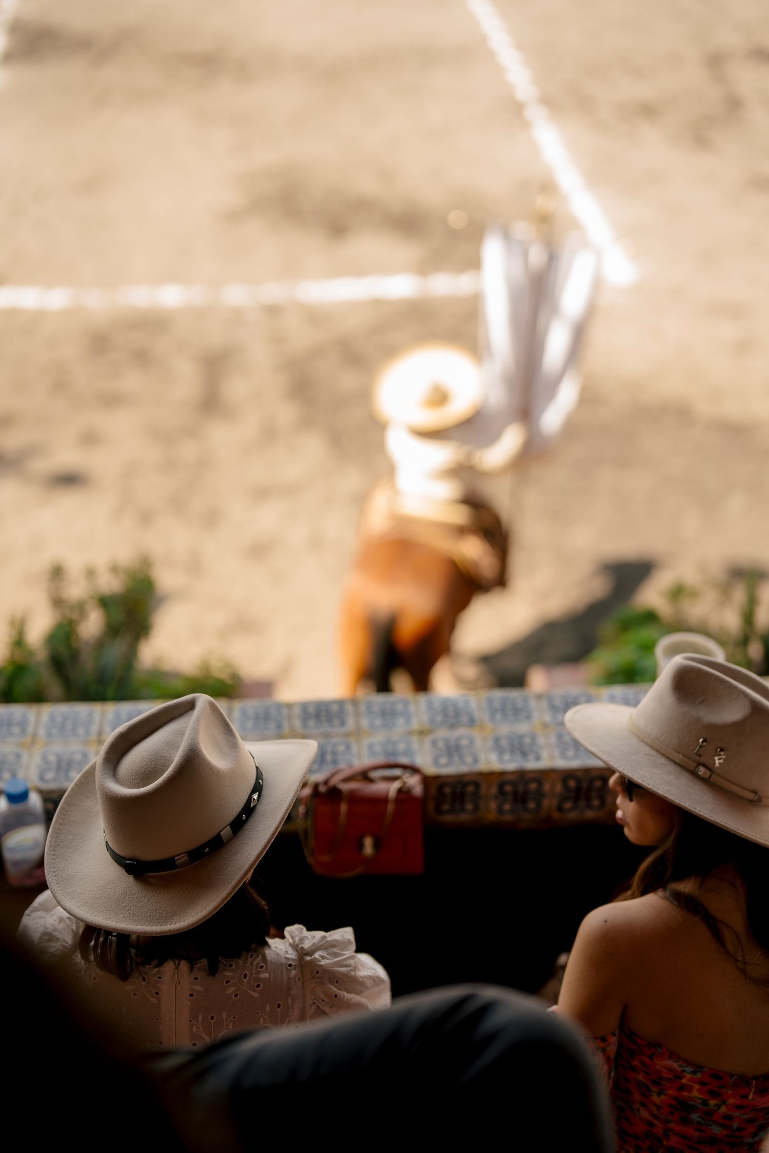 Two women in cowboy hats watch a horse rider perform in a sandy arena at a traditional rodeo event.