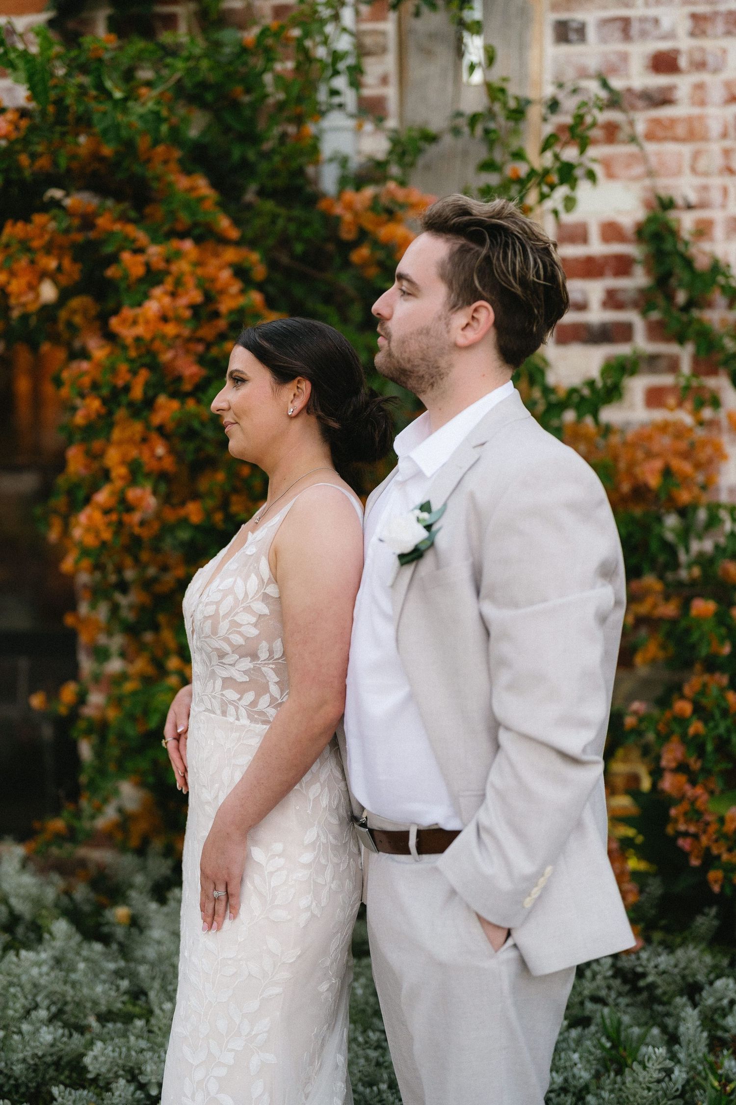 Bride in lace gown and groom in tan suit pose together before orange bougainvillea and brick wall backdrop.