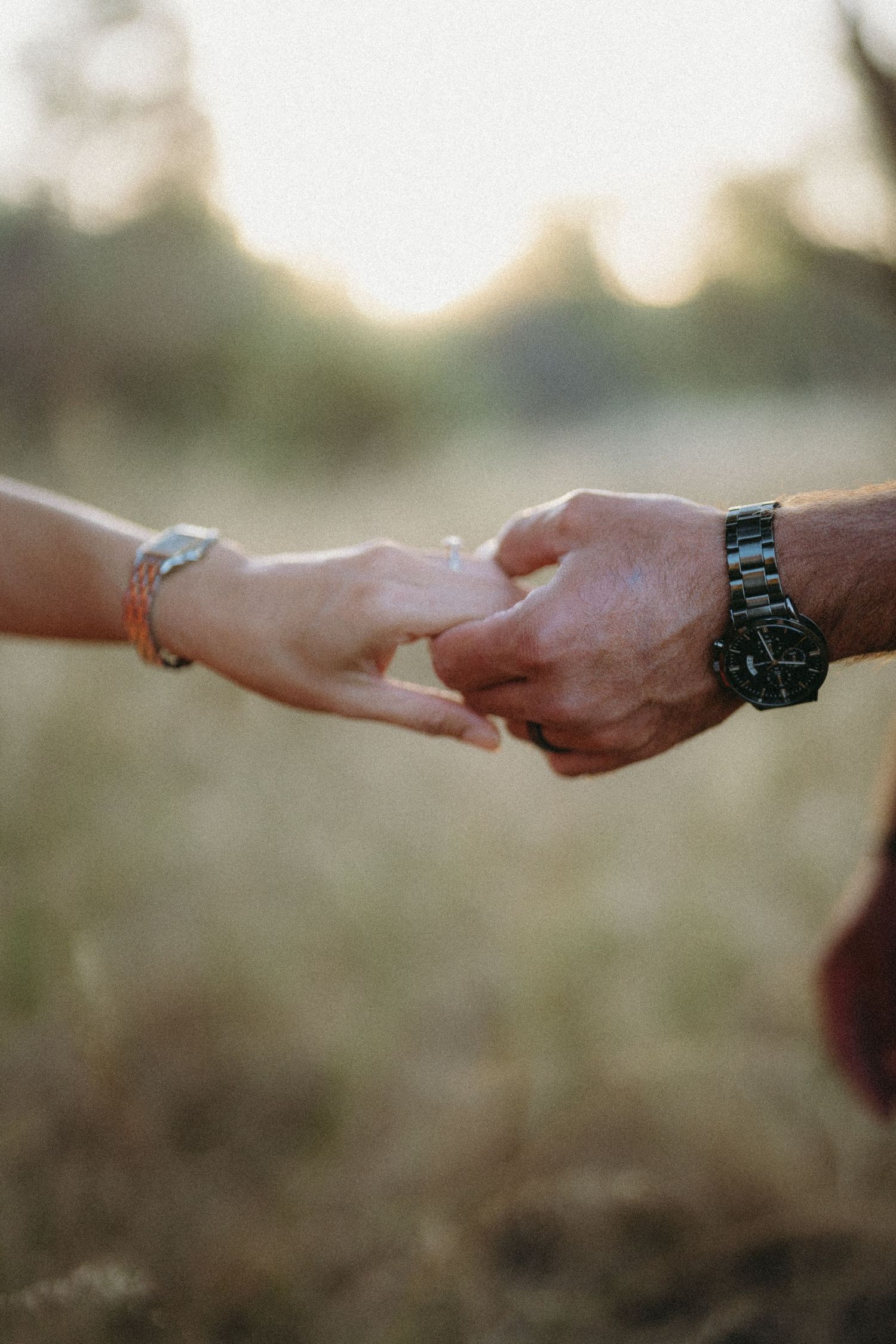 Two people gently holding hands outdoors in a golden field, one wearing a bracelet and the other a black watch.
