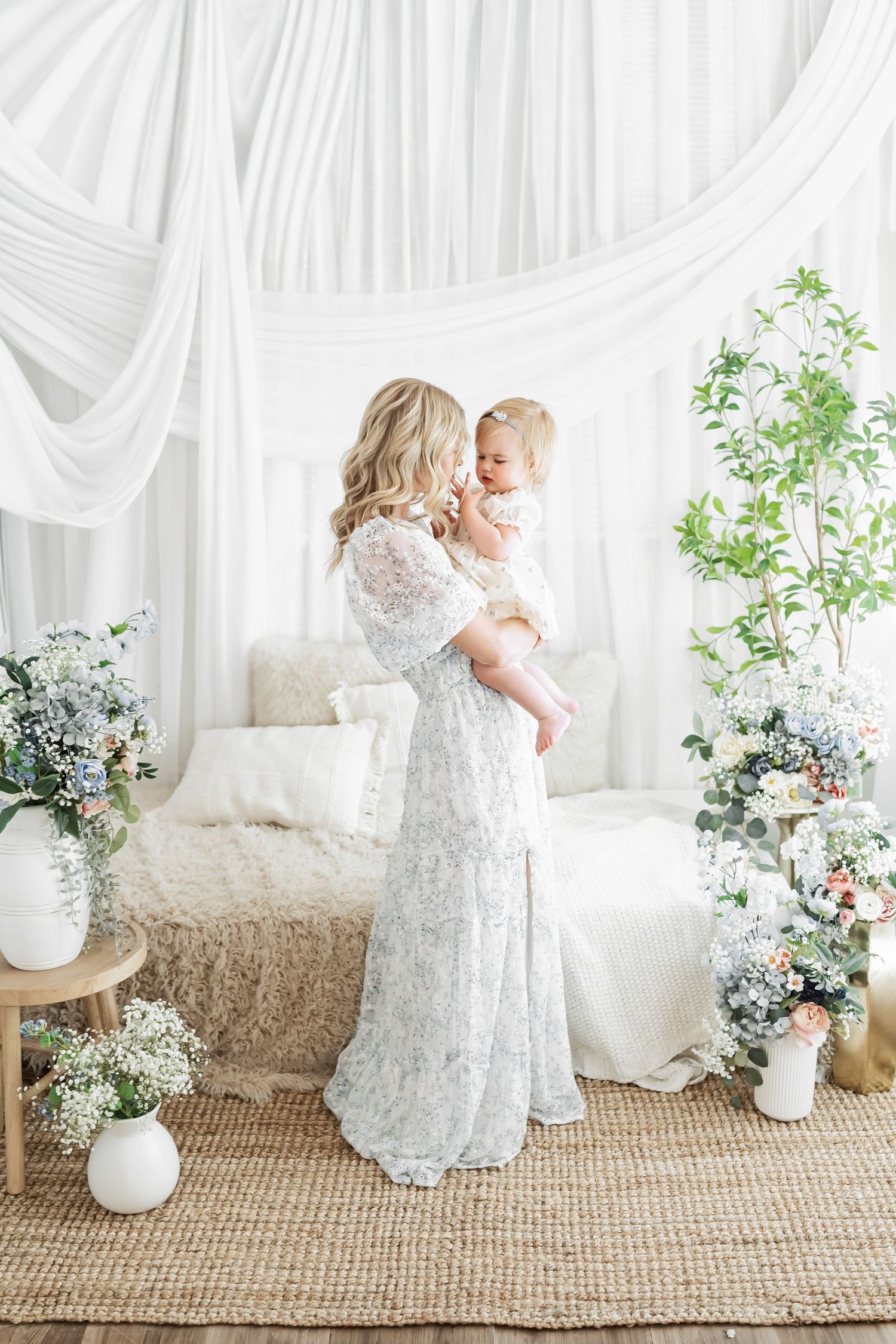 A woman in a white lace gown holds a baby in a floral-decorated studio with white drapes and greenery.