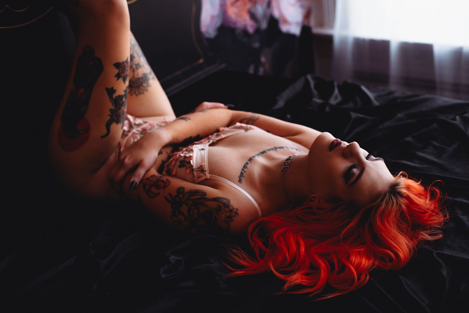 Woman with red hair and tattoos posing in lingerie on dark bedding in a moody boudoir photography setting.