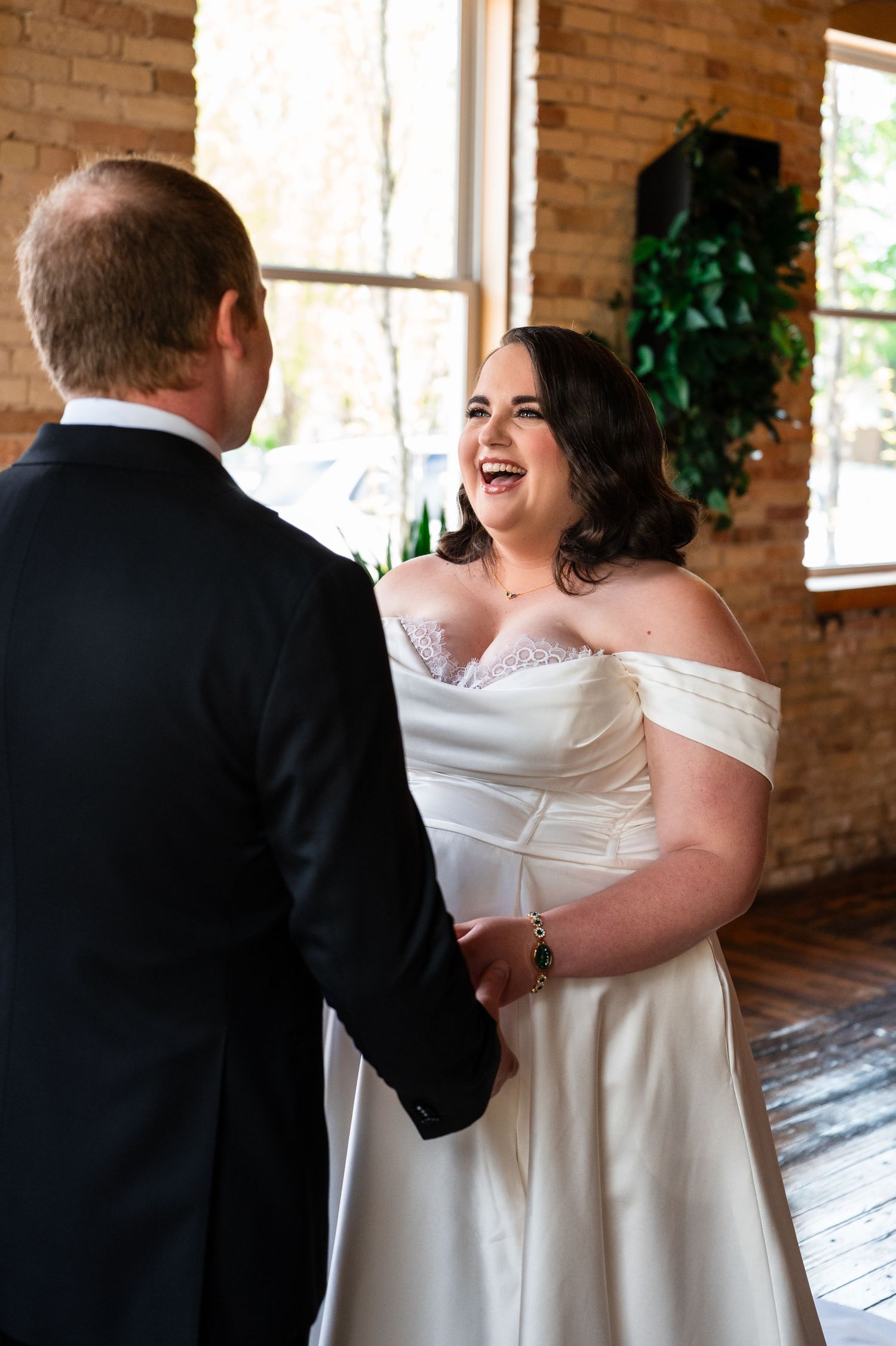 Joyful bride in off-shoulder white gown laughing with groom in black tuxedo during wedding first look.