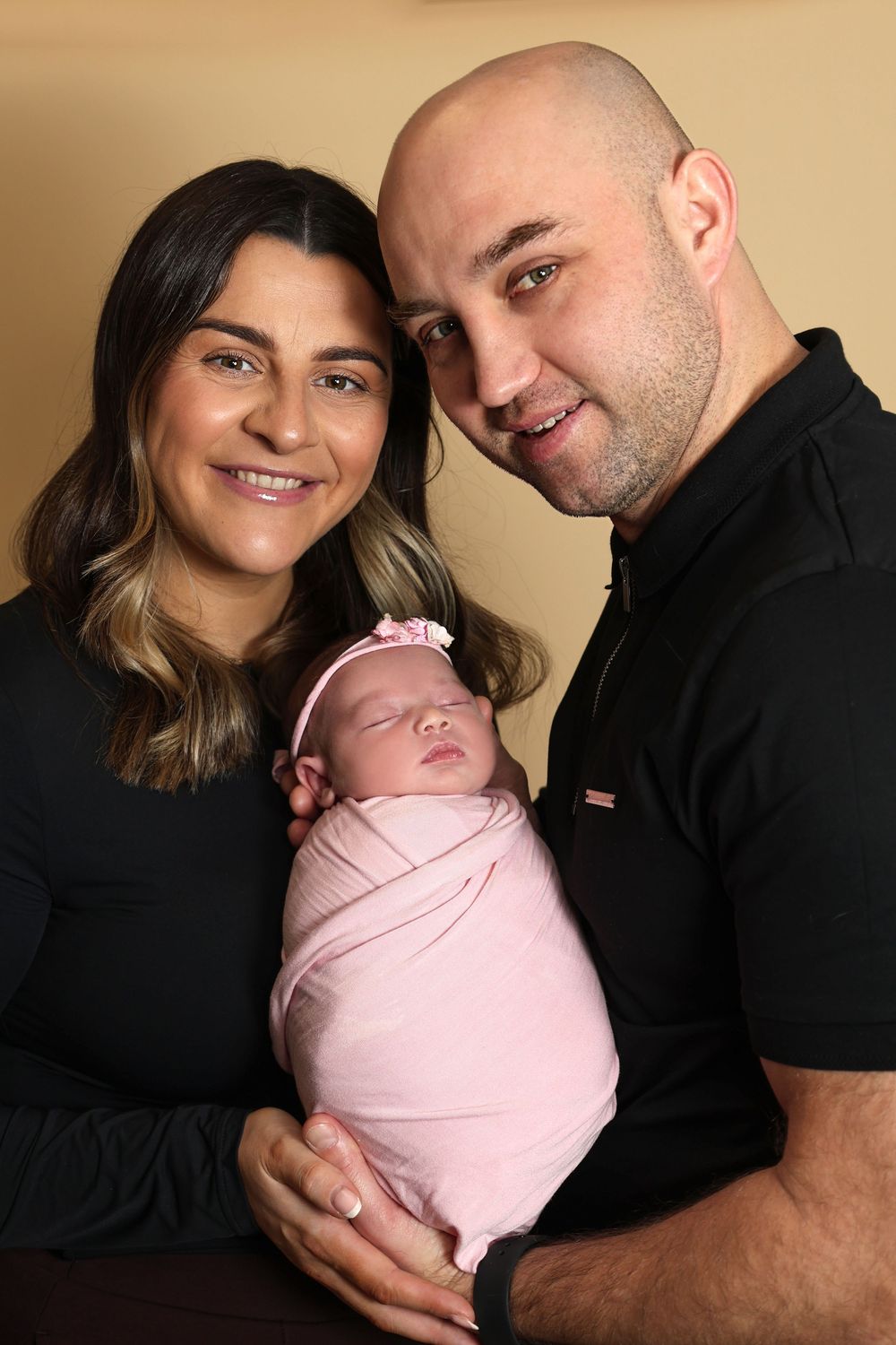 Happy couple in black outfits smiling while holding their sleeping newborn baby wrapped in pink.