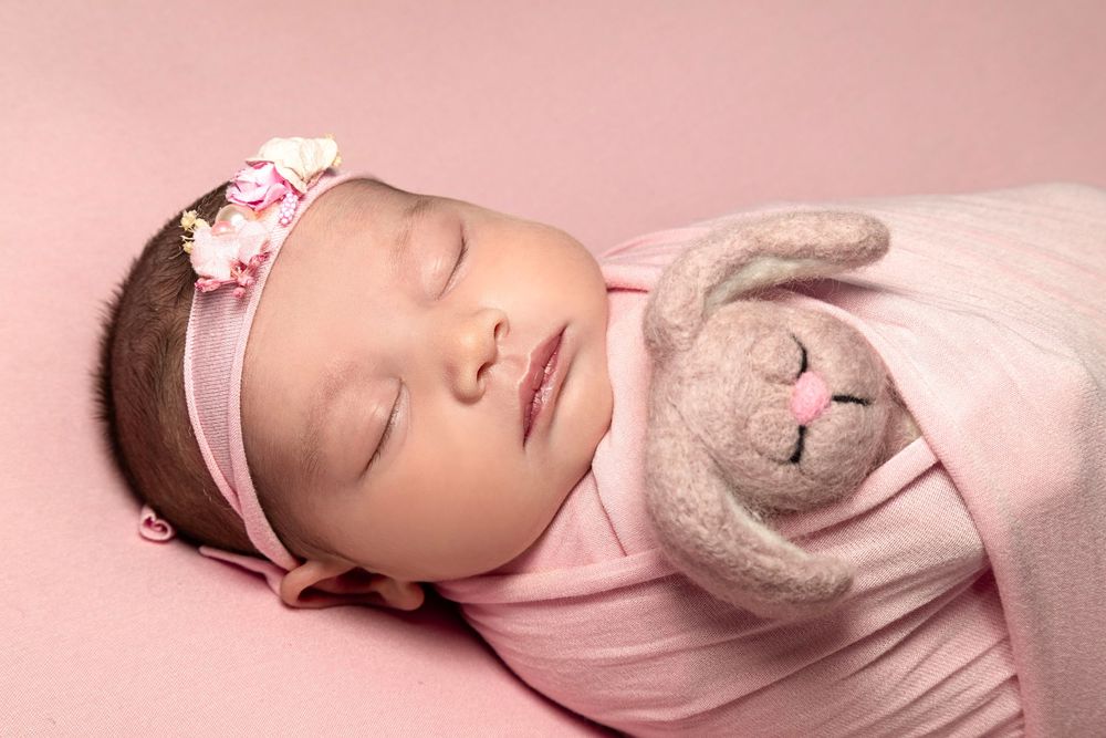 Newborn baby girl sleeping peacefully wrapped in pink, wearing a floral headband, cuddling a soft bunny toy.