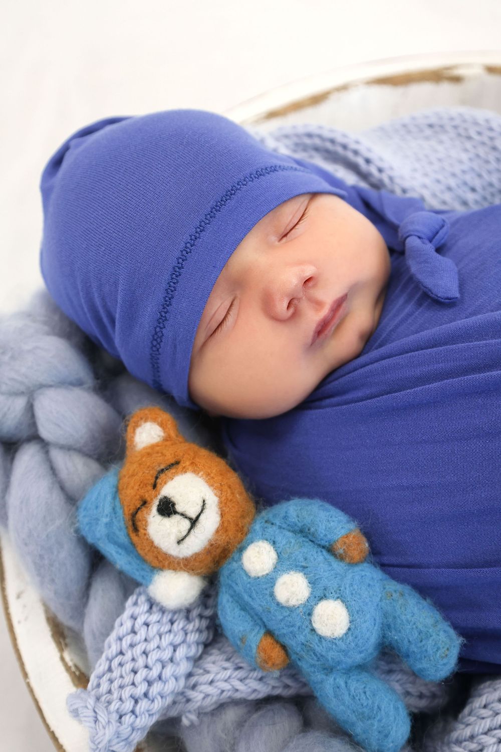 Newborn baby sleeping in a basket, wrapped in blue swaddle with matching hat, cuddling a small felted teddy bear toy.