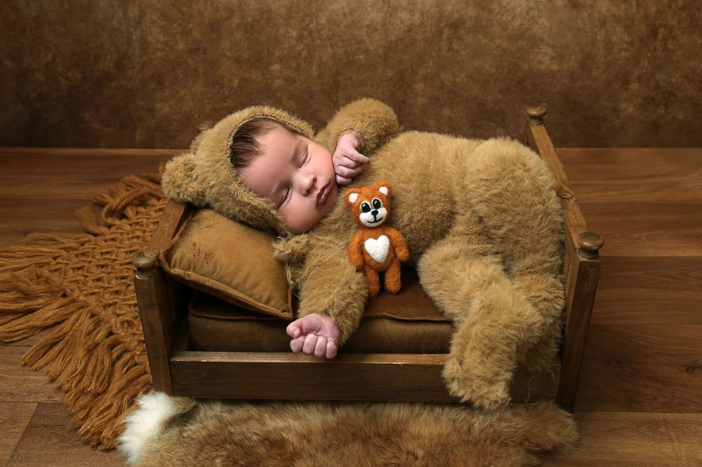 Newborn baby in brown bear outfit sleeping on tiny wooden bed with matching teddy bear toy.