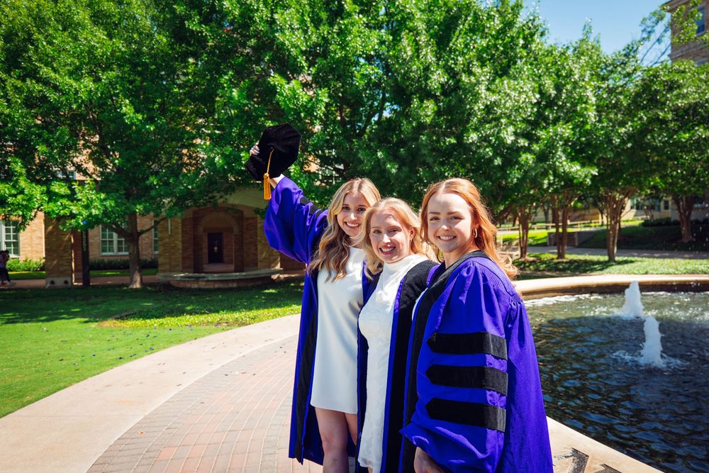 Three women in graduation regalia smile and pose near a fountain on a sunny college campus.