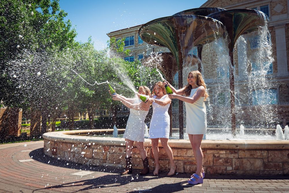 Three women in white dresses celebrate by spraying champagne near a decorative fountain outdoors.