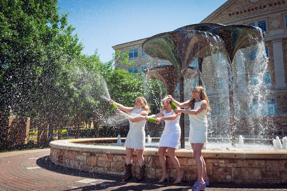 Three women in white dresses celebrating by a decorative fountain, splashing water outdoors.