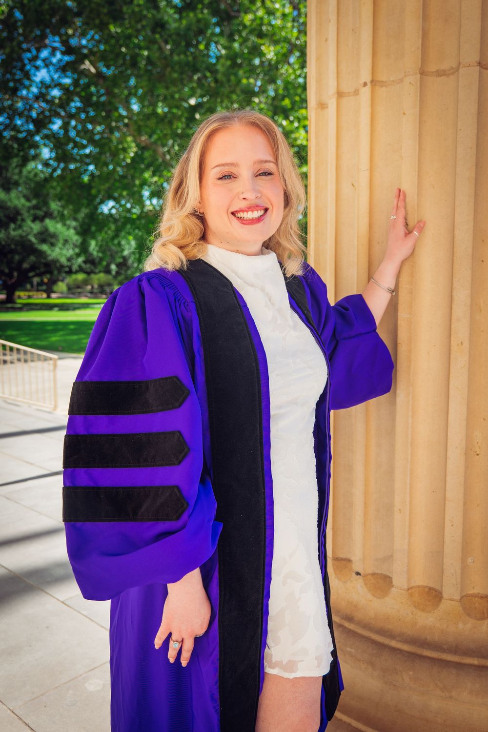 Smiling blonde woman in purple doctoral graduation gown posing by stone columns outdoors.