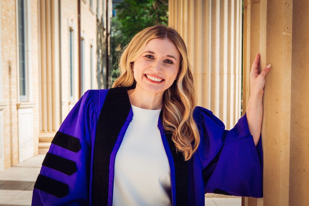 Smiling woman in blue graduation robe poses between columns outdoors for doctoral degree ceremony photo.