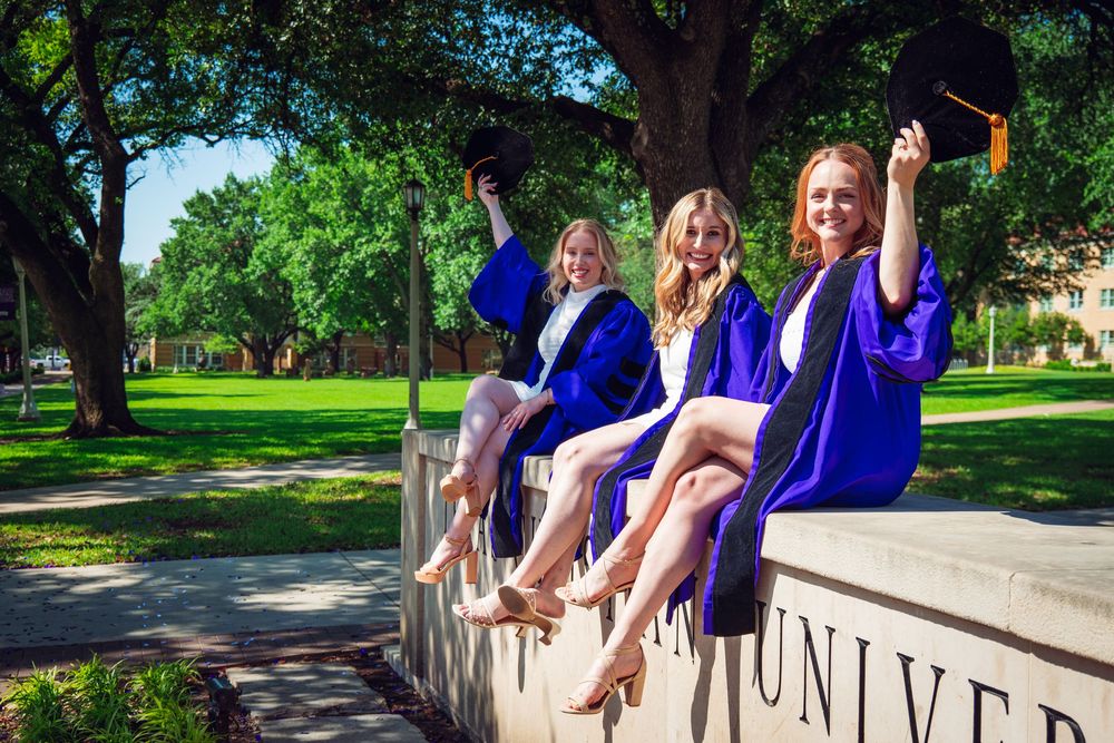 Three smiling women in blue graduation gowns sit on a university wall, holding caps outdoors.