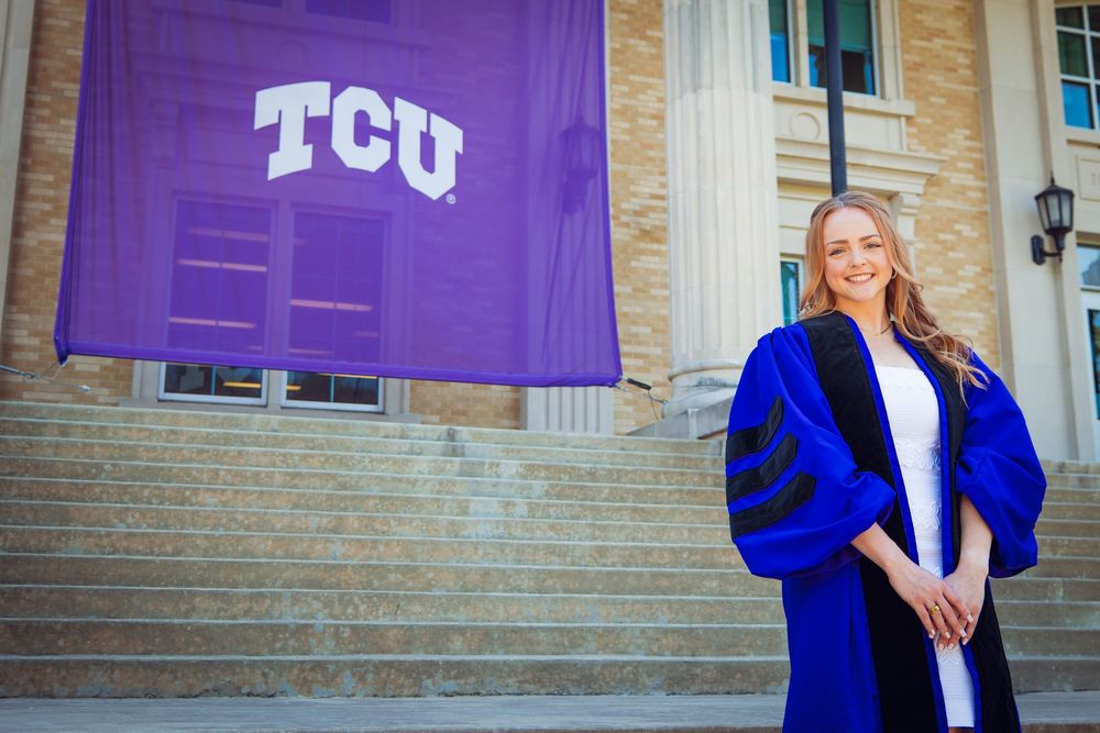 TCU graduate smiles in blue doctoral regalia on steps before purple TCU banner at Texas Christian University.