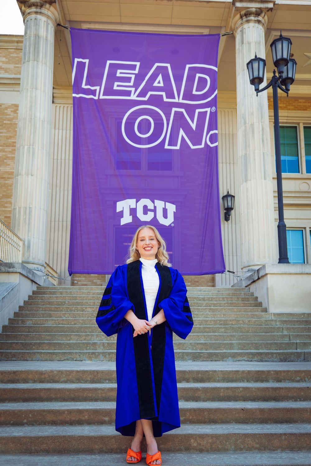 Woman in blue doctoral regalia smiles on steps before a purple TCU 'Lead On' banner.