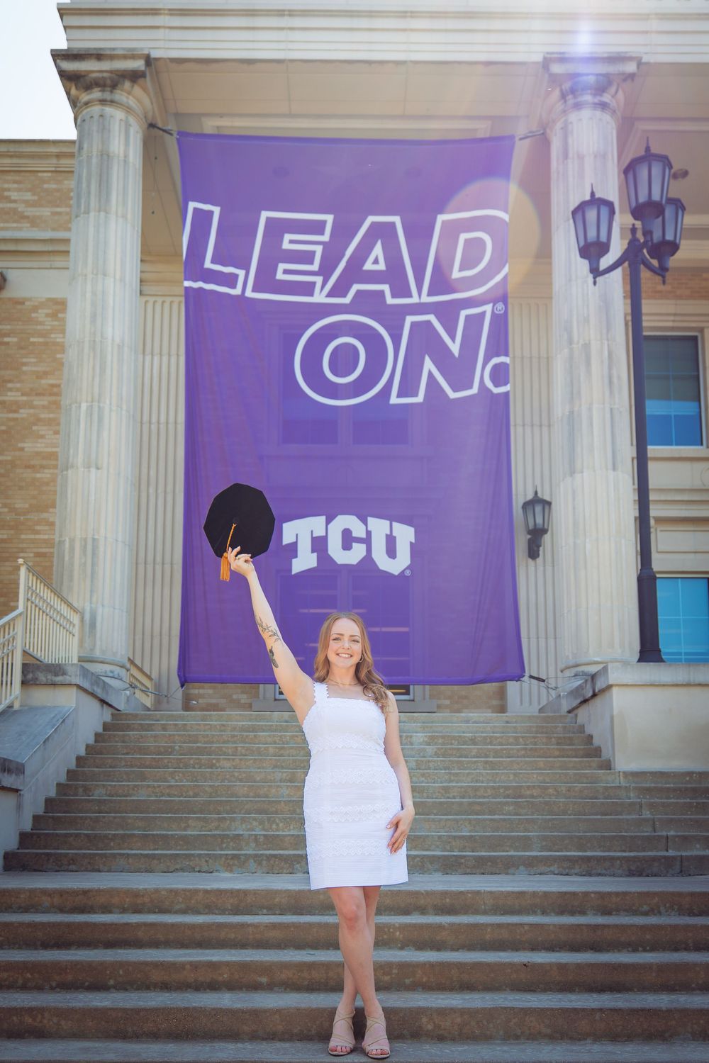 TCU graduate in white dress celebrates on steps holding cap in front of purple Lead On banner.