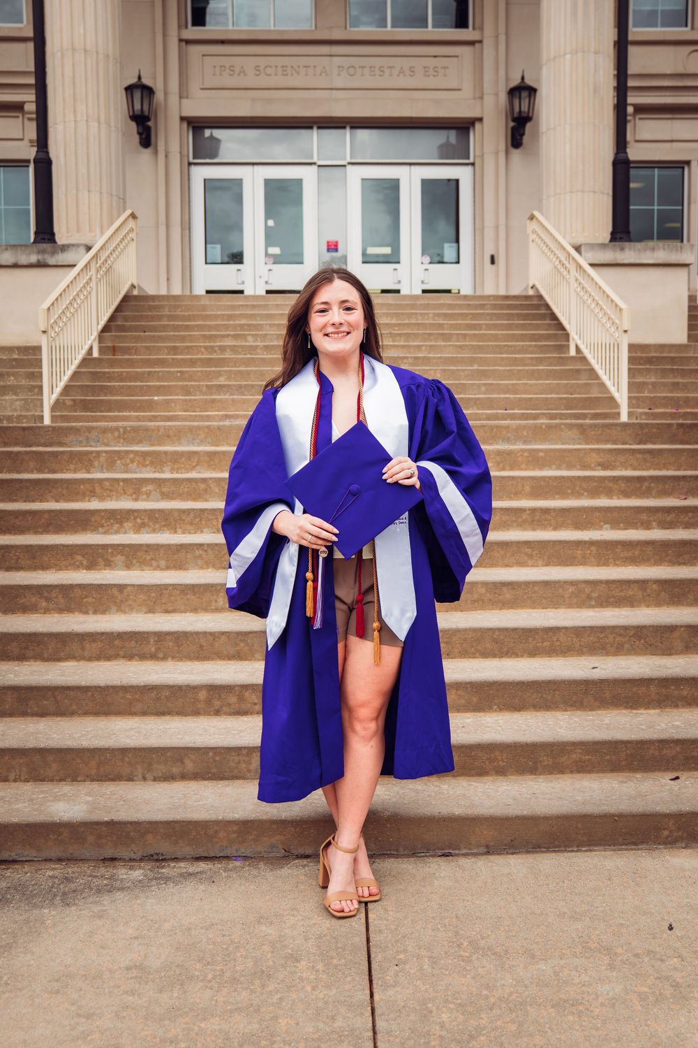 Young woman in purple graduation gown and cap smiling on university steps during commencement celebration.