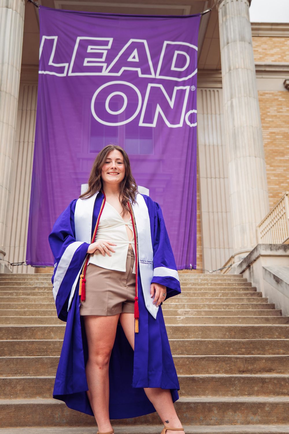 Young woman in purple graduation gown smiling on steps before a purple Lead On banner at university commencement.