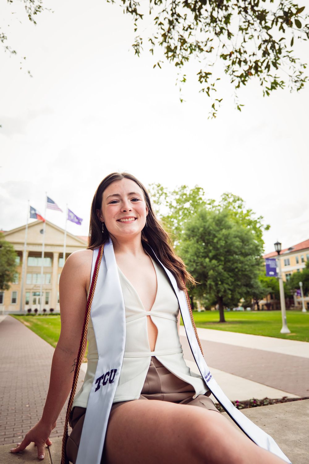 Smiling TCU graduate wearing white sash sits outdoors on campus with flags and green trees in background.
