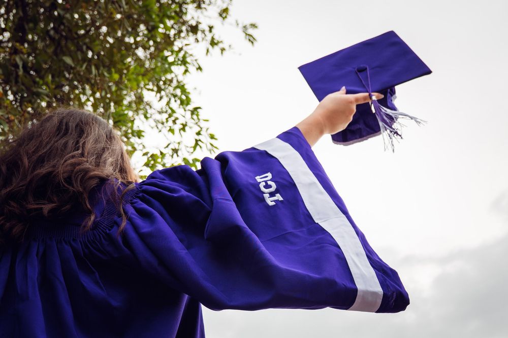 A graduate in purple cap and gown raises their mortarboard triumphantly against a bright sky with green trees.