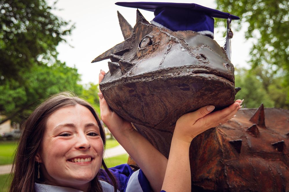 Smiling graduate poses with a bronze horned frog statue wearing a blue graduation cap outdoors.