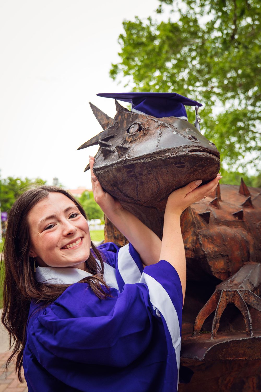 Smiling graduate in purple gown places graduation cap on bronze horned frog statue outdoors.