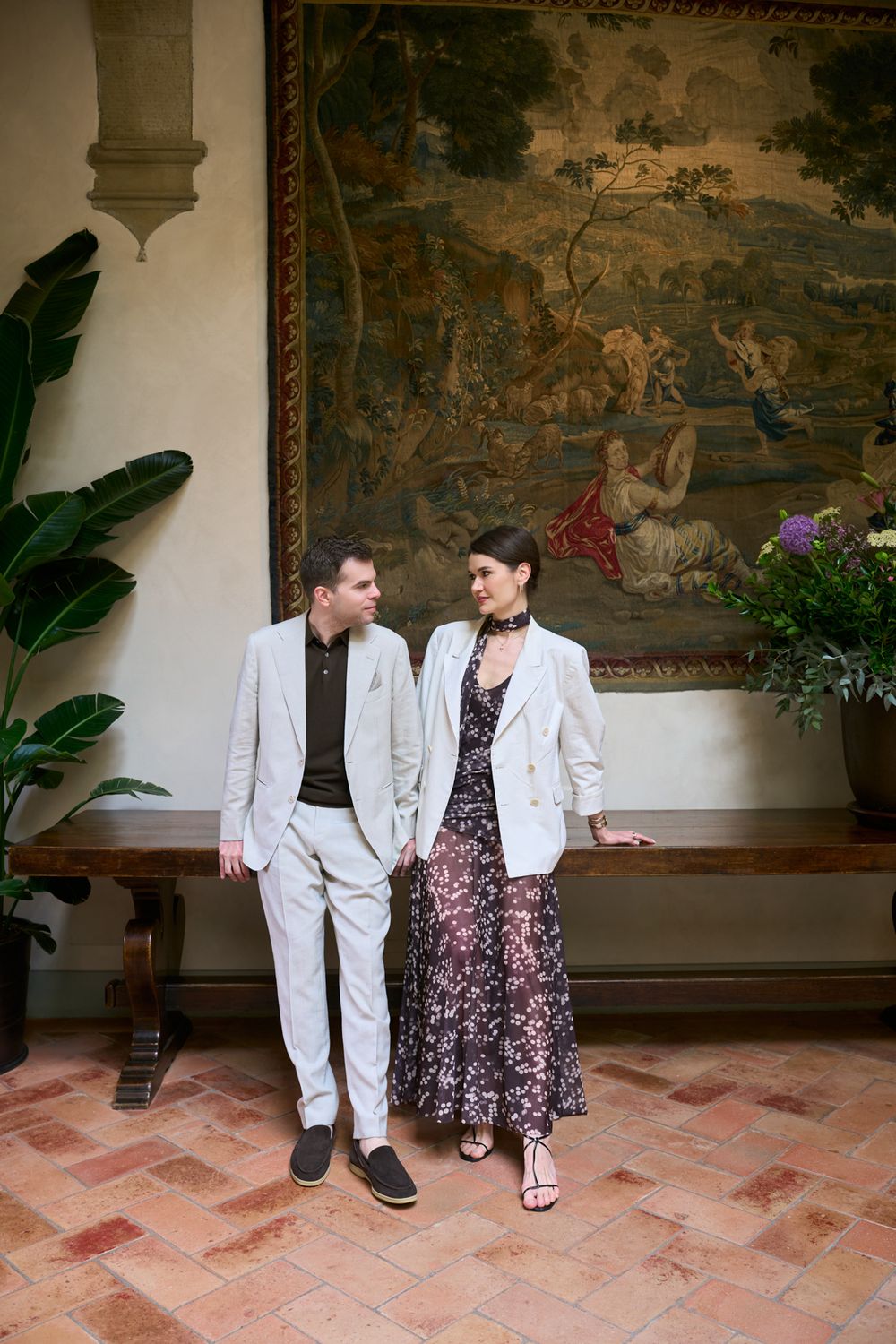 Couple in elegant attire posing before a large tapestry in a grand hall with terracotta floors.