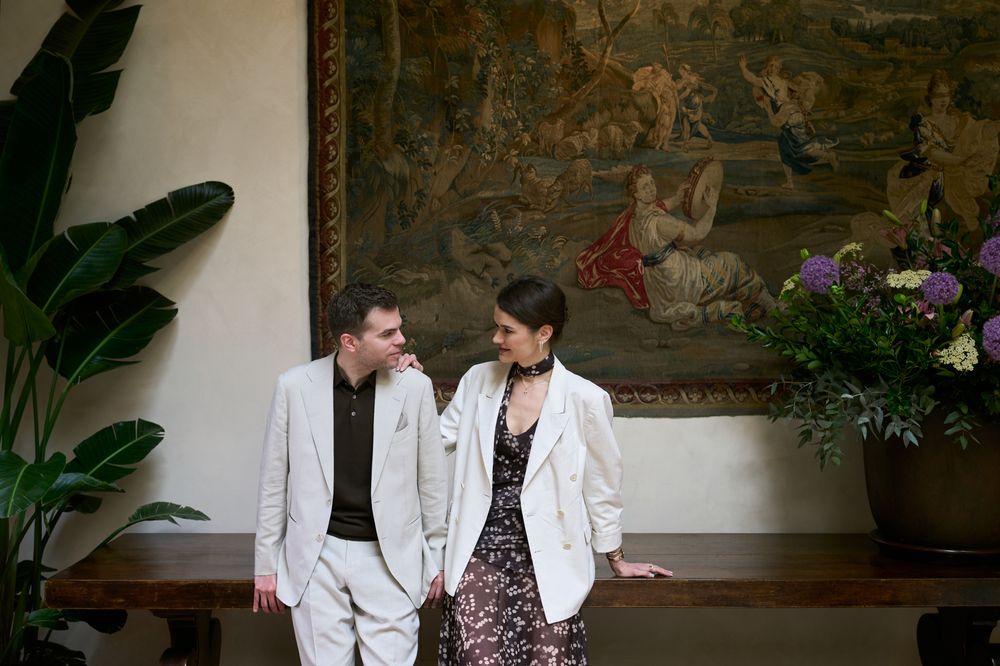 Elegant couple in white blazers posing together indoors before a grand tapestry with floral arrangements.