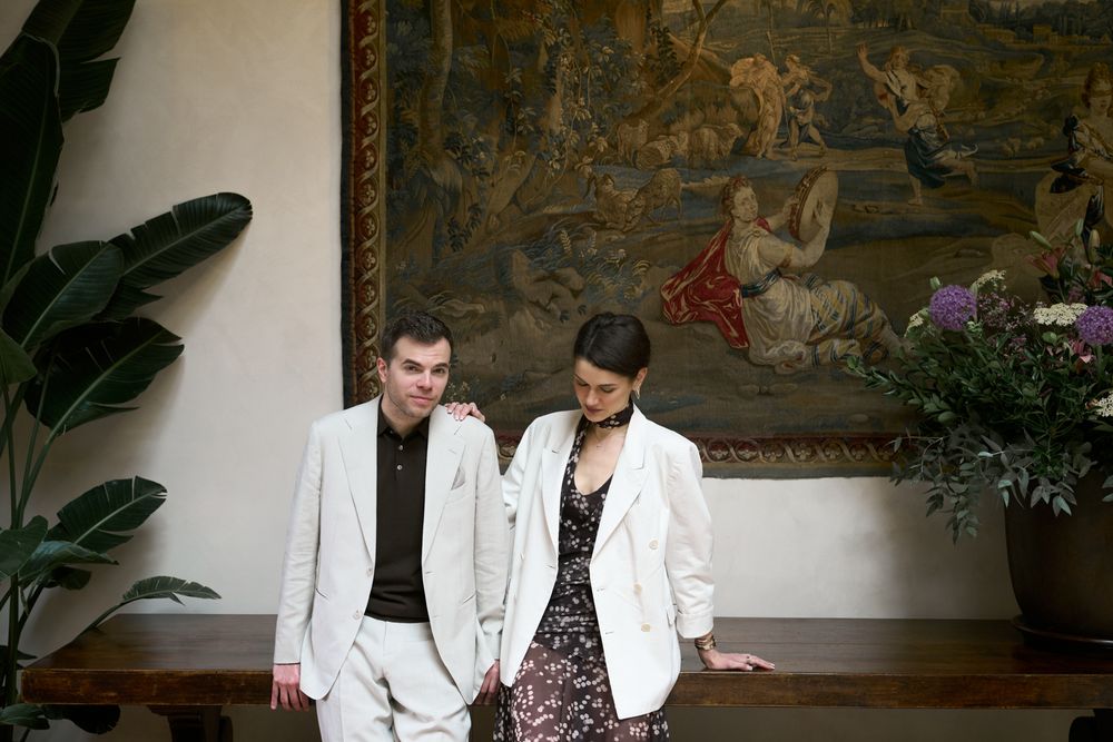 A man and woman in elegant attire stand together before a large ornate tapestry in a sophisticated interior.