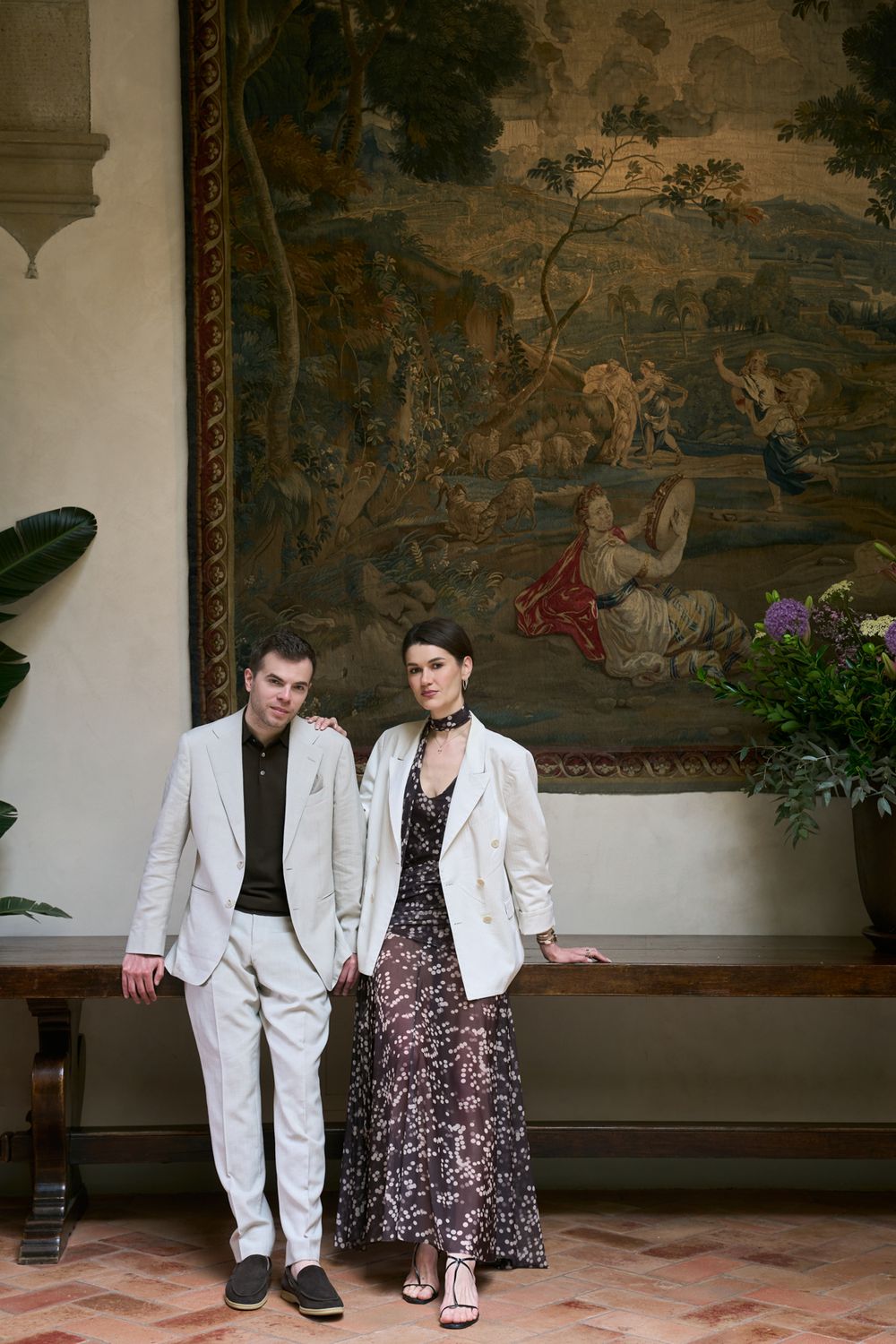 A couple poses elegantly before a grand tapestry, wearing a white suit and floral maxi dress with blazer in a classic interior.