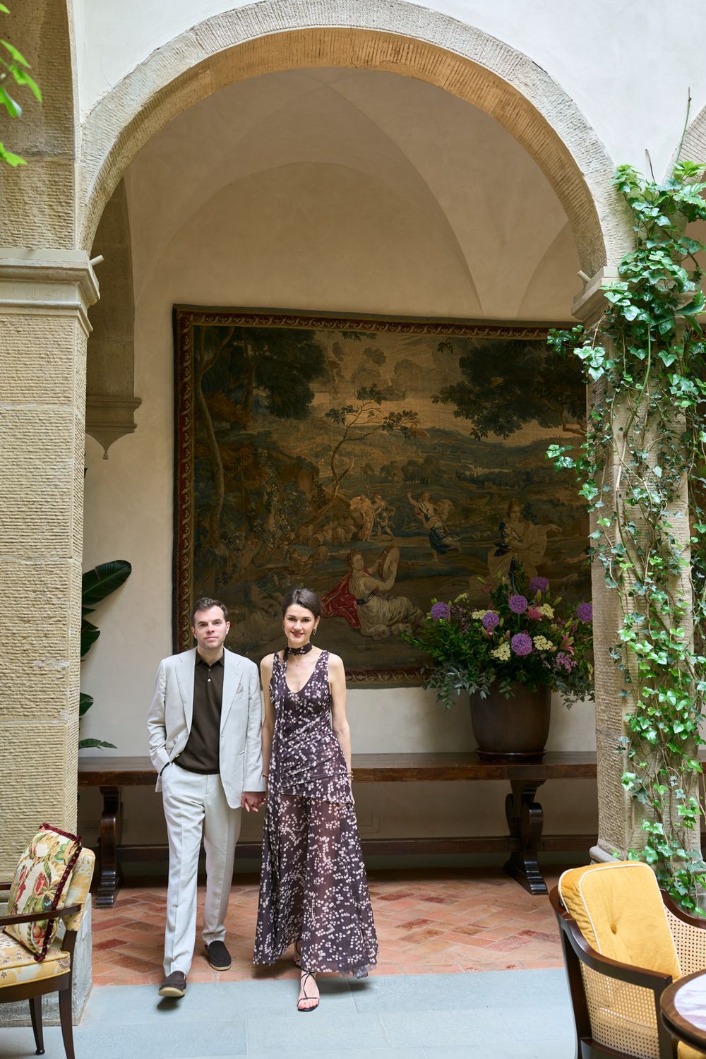 Elegant couple standing under stone arch before ornate tapestry in a luxurious villa courtyard with ivy and flowers.