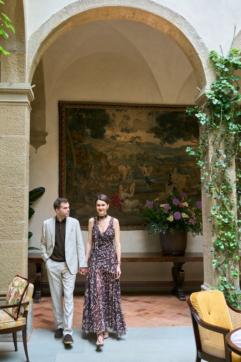 Elegant couple holding hands walking through arched stone hallway with tapestry and ivy decor.
