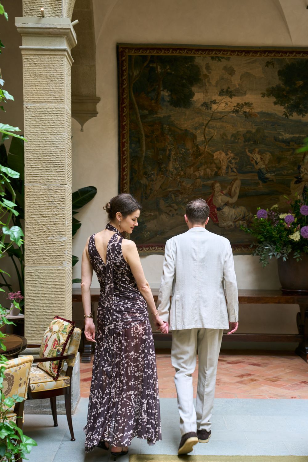 Couple holding hands walking toward a large tapestry in an elegant indoor courtyard, woman in floral dress, man in beige suit.