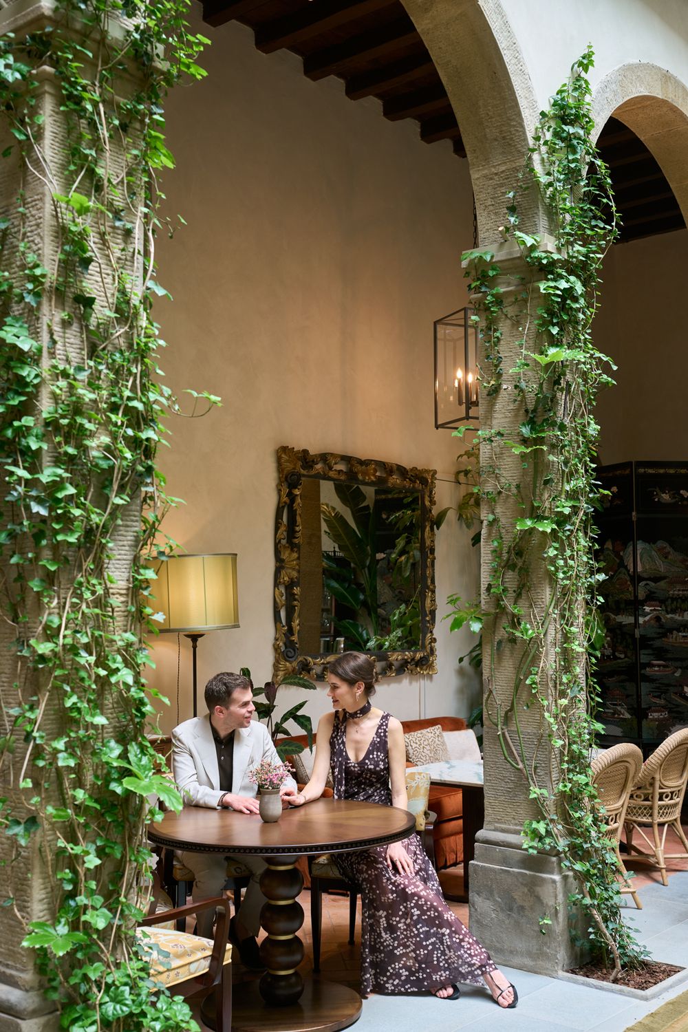 Couple sitting at a round wooden table in an elegant courtyard with ivy-draped stone arches and warm ambient lighting.