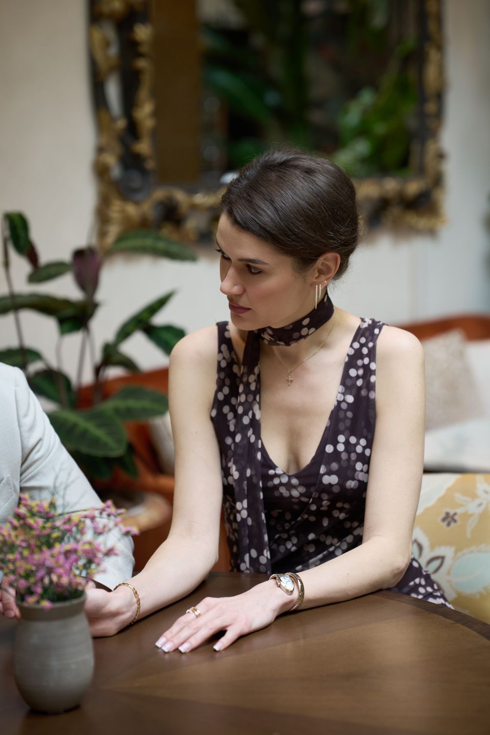 Young woman in floral dress with neck scarf sits at wooden table, plants and gold mirror in background.