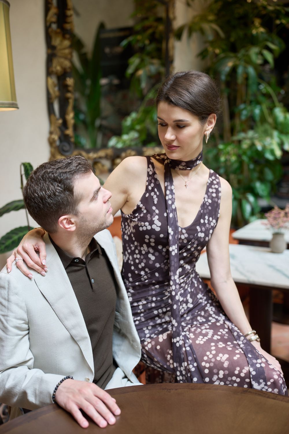 Elegant couple sharing an intimate moment at a table, woman in floral dress with hand on mans shoulder in lush interior.