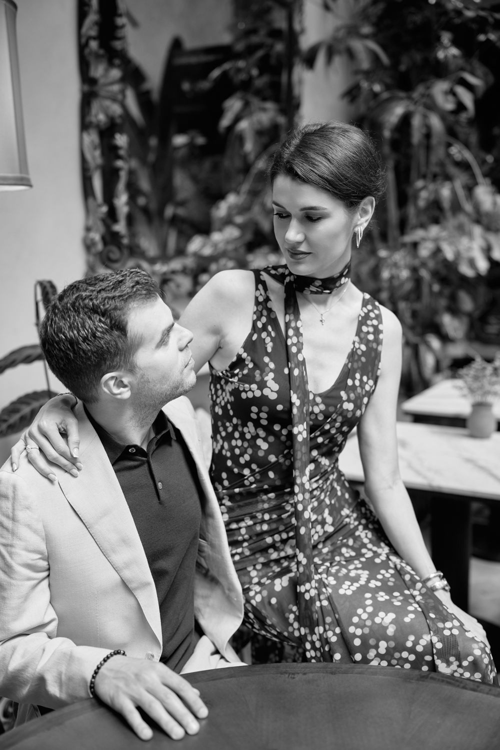Black and white photo of an elegant couple sharing an intimate moment at a restaurant table surrounded by lush plants.