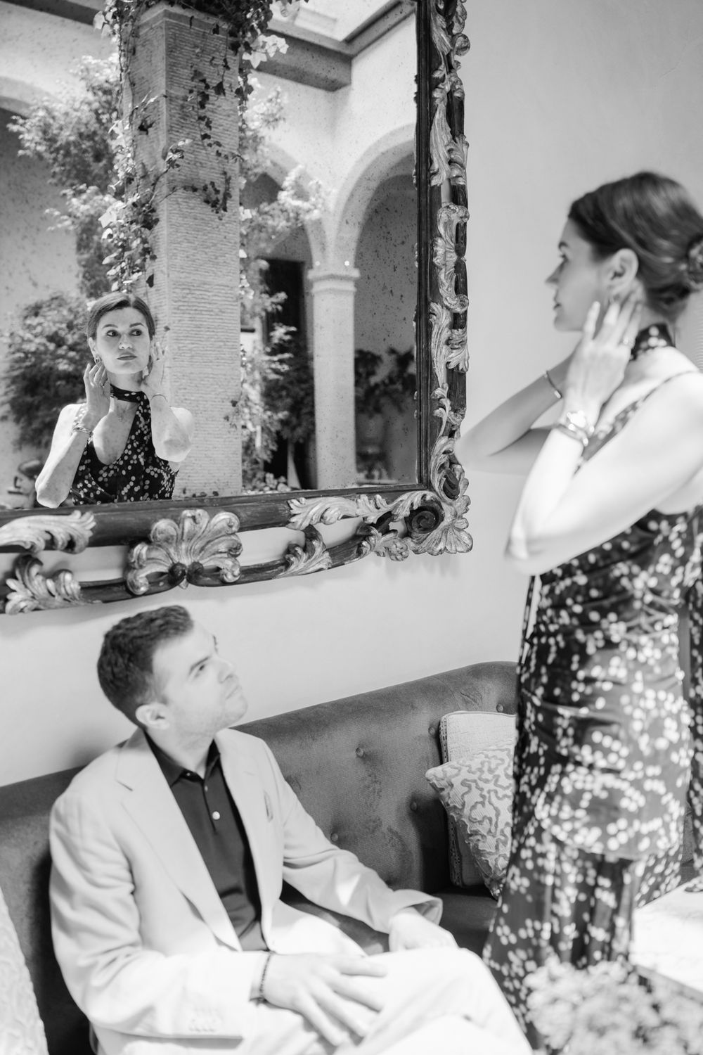 Black and white photo of a man seated on a sofa watching a woman adjust her jewelry in an ornate mirror.